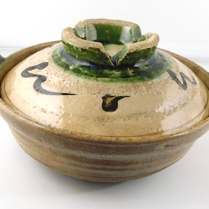May include: A round ceramic cooking pot with a lid, featuring a beige base color with dark green glazed accents on the handle and lid knob. The lid is decorated with hand-painted black brushstroke markings and has two side handles for lifting.