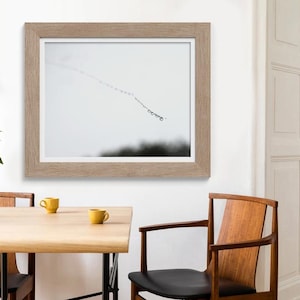 May include: A framed monochrome print showing a line of small, dark shapes against a light background. The print is in a light brown wooden frame and hangs on a white wall. The room also features a wooden table and chairs.