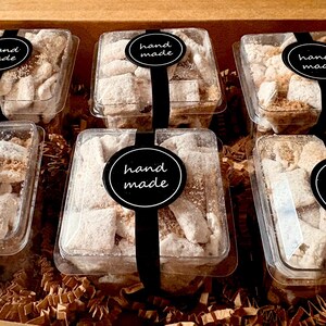 May include: Six clear plastic containers filled with a sweet treat, each sealed with a black ribbon and a "hand made" sticker. The treats are light-colored, possibly dusted with powdered sugar, and sit in a cardboard box with brown paper filler.