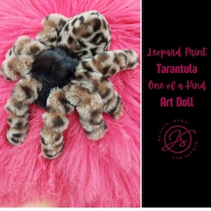 May include: A leopard print tarantula art doll with a black body and brown and tan legs, set against a vibrant pink background. The doll is made of soft, plush material. Text on the right side reads "Leopard Print Tarantula One of a Kind Art Doll".
