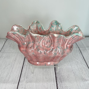 May include: A pink and green ceramic planter with a scalloped edge. The planter has a unique, textured surface and is shaped like a seashell.