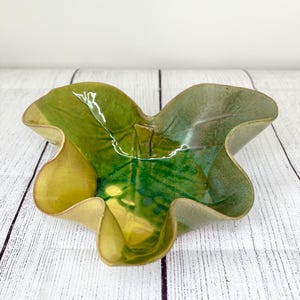 May include: A handcrafted ceramic bowl in the shape of a leaf. The bowl features a glossy finish with shades of green and yellow. The edges are gently ruffled, adding a unique touch to this decorative piece.