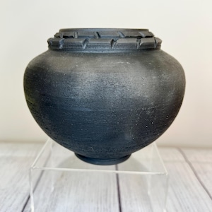 May include: A dark gray ceramic vase with a textured surface. The vase has a rounded body and a decorative rim. It is displayed on a clear acrylic stand. The vase is approximately 8 inches tall.