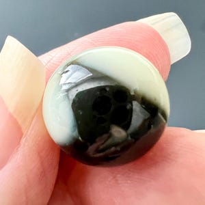 May include: A small, spherical, polished stone bead featuring a smooth, glossy surface with a distinct color split. One side is a creamy, pale off-white, while the other side is a deep, solid black with visible natural surface indentations.