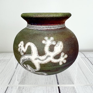 May include: A green ceramic vase with a white lizard design. The vase has a rounded body and a slightly flared rim.