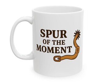 May include: White ceramic mug with the text "SPUR OF THE MOMENT" in brown, accompanied by a brown spur illustration. The mug has a curved handle and is suitable for hot or cold beverages.
