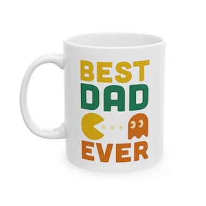 May include: White ceramic mug with the words "BEST DAD EVER" in yellow, green, and orange. The design includes Pac-Man and a ghost character. The mug has a curved handle.