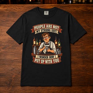 May include: A black short-sleeved t-shirt featuring a graphic of a bartender wiping a glass. The text reads, "PEOPLE ARE NOT MY STRONG SUIT, I WONDER HOW I PUT UP WITH YOU." The design includes bottles, a shaker, and a jigger on a wooden bar counter.
