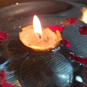 K&ouml;nnte beinhalten: Eine kleine Kerze in einer Walnussschale mit brennender Flamme, die in einer mit Wasser gef&uuml;llten, klaren Glasschale schwimmt. Mehrere dunkelrote Rosenbl&auml;tter sind auf der Wasseroberfl&auml;che verstreut und umgeben die schwimmende Walnusskerze.