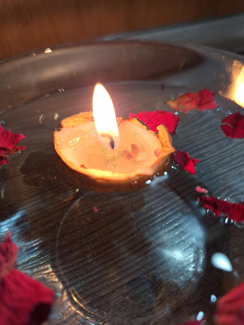 K&ouml;nnte beinhalten: Eine kleine Kerze in einer Walnussschale mit brennender Flamme, die in einer mit Wasser gef&uuml;llten, klaren Glasschale schwimmt. Mehrere dunkelrote Rosenbl&auml;tter sind auf der Wasseroberfl&auml;che verstreut und umgeben die schwimmende Walnusskerze.