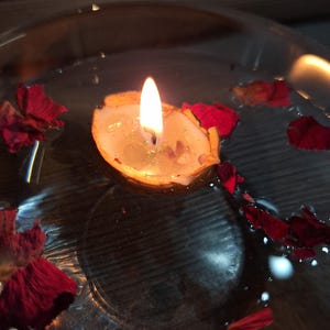K&ouml;nnte beinhalten: Eine kleine Kerze in einer Walnussschale, die in einer mit Wasser gef&uuml;llten klaren Glasschale schwimmt. Die Kerzenflamme brennt und verbreitet einen warmen Schein. Mehrere dunkelrote Rosenbl&auml;tter schwimmen auf der Wasseroberfl&auml;che.
