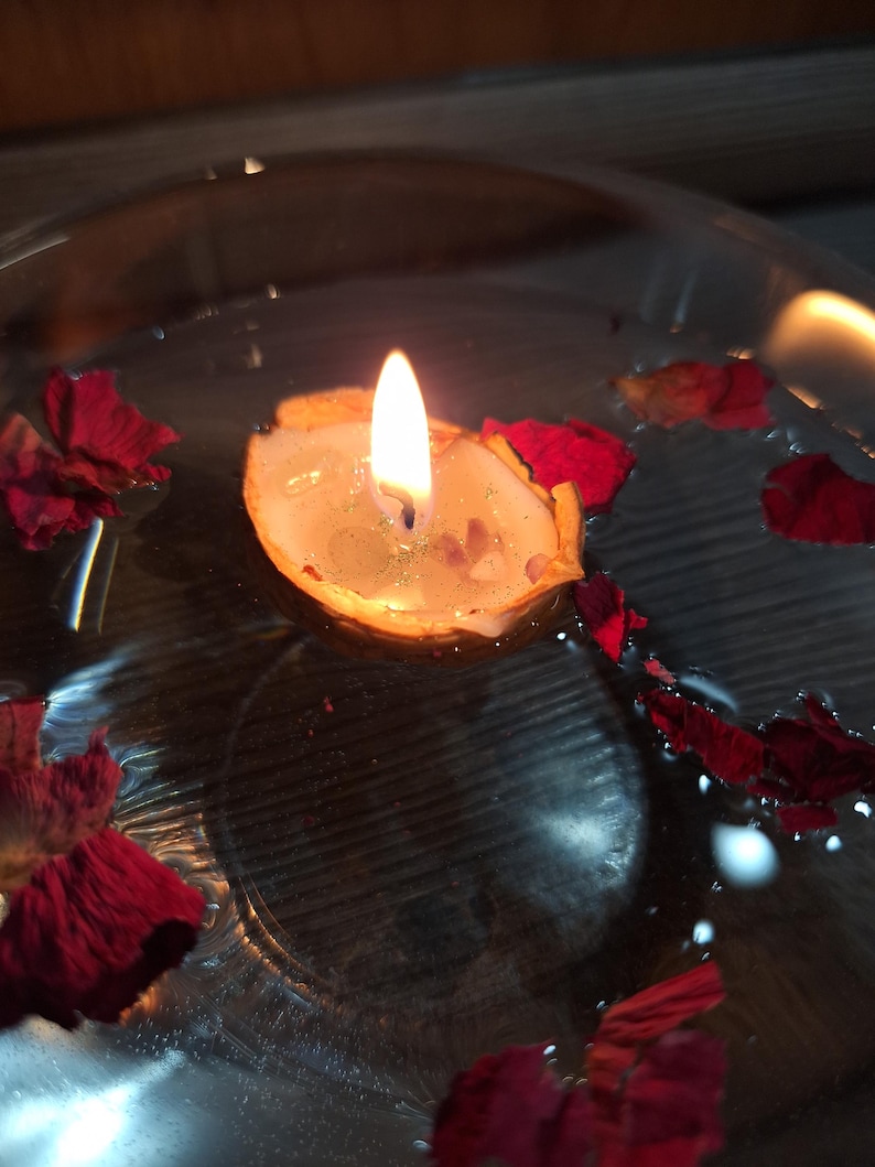 K&ouml;nnte beinhalten: Eine kleine Kerze in einer Walnussschale, die in einer mit Wasser gef&uuml;llten klaren Glasschale schwimmt. Die Kerzenflamme brennt und verbreitet einen warmen Schein. Mehrere dunkelrote Rosenbl&auml;tter schwimmen auf der Wasseroberfl&auml;che.