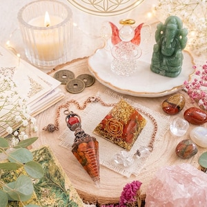 Könnte beinhalten: Eine Sammlung spiritueller Gegenstände, darunter ein Pendel aus rotem und goldenem Harz mit Kupferkette, eine Pyramide aus rotem und goldenem Harz mit Wurzelchakra-Symbol, eine Glasengel-Figur, eine grüne Ganesha-Statue und ein Rosenquarz.