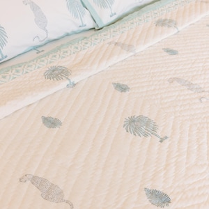May include: A cream-colored quilt and pillow set with a repeating pattern of light blue palm trees and leopards. The bedding has a light blue border and is quilted with a diamond pattern. The pillowcases feature the same design.