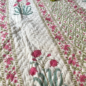 May include: White cotton quilt with a floral pattern in shades of pink, green, and blue. The quilt has a quilted texture and is decorated with a repeating pattern of flowers and leaves.