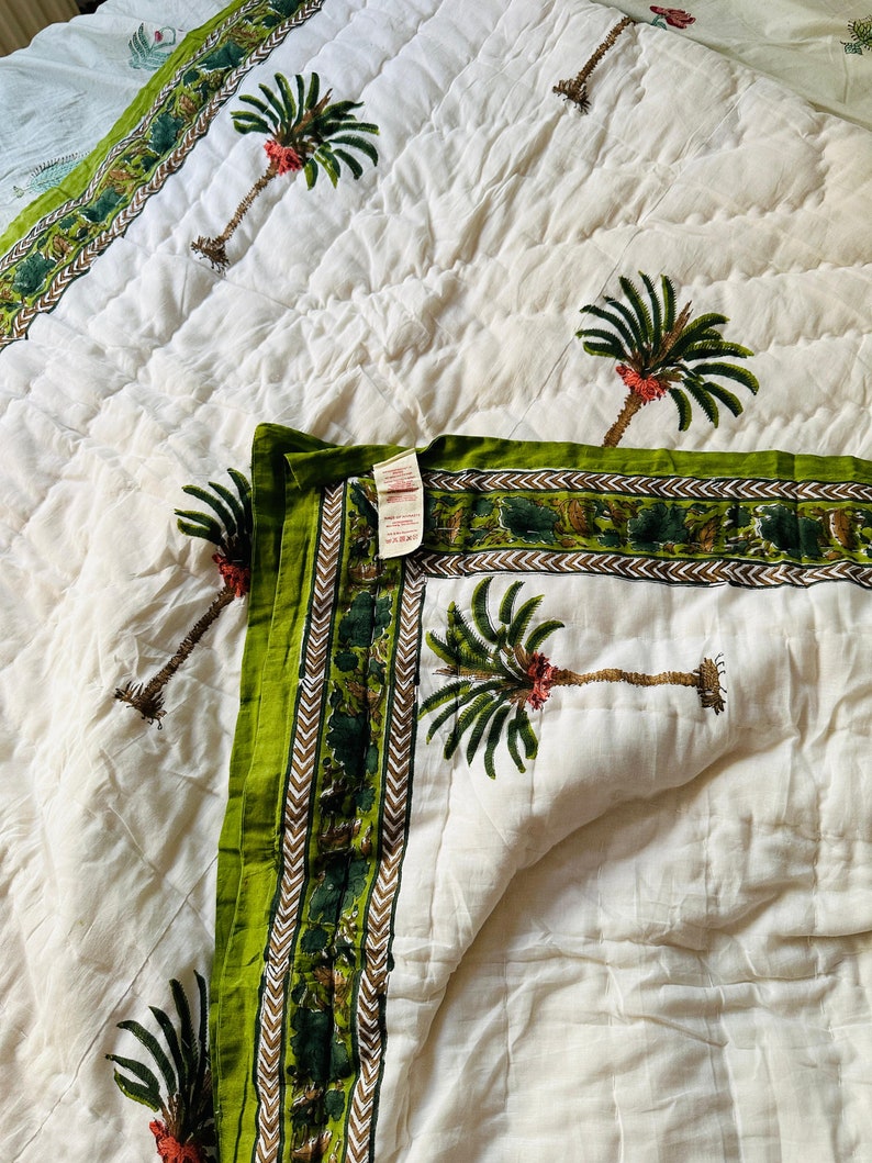 May include: White quilted bedspread with a green border and palm tree design. The border features a repeating pattern of green and brown elements. The palm trees have green fronds and red accents. The bedspread is folded, showing the pattern.