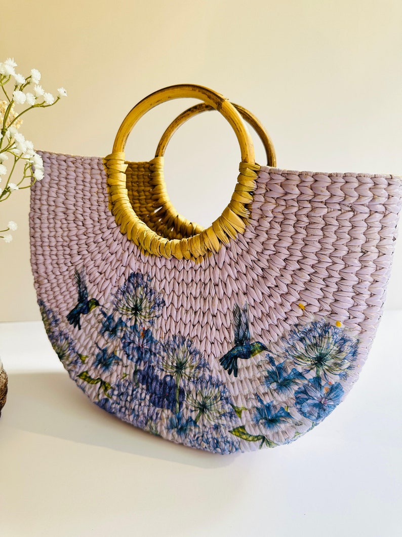 May include: A lavender woven tote bag with a floral print and two brown wooden handles. The print features blue hummingbirds and flowers.
