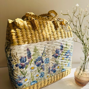 Puede incluir: Un bolso de mano de paja tejido con estampado floral. El bolso tiene un fondo blanco con flores azules y moradas. El bolso est&aacute; hecho de materiales naturales y tiene un aspecto r&uacute;stico.