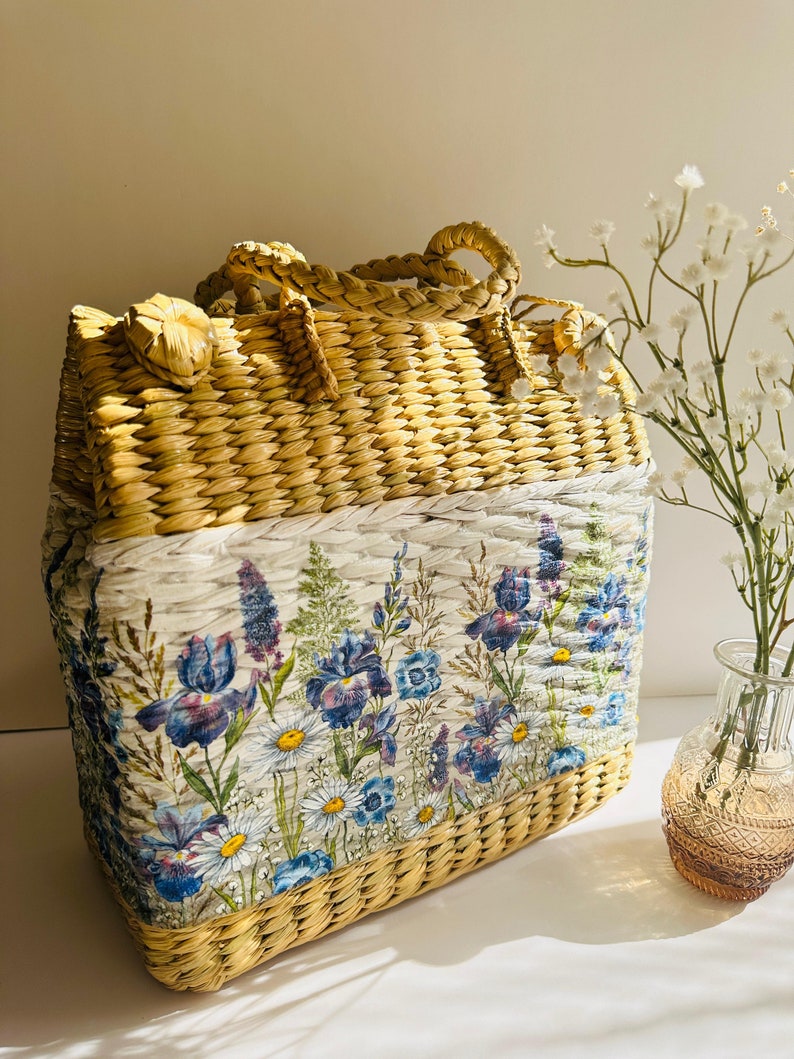 Puede incluir: Un bolso de mano de paja tejido con estampado floral. El bolso tiene un fondo blanco con flores azules y moradas. El bolso est&aacute; hecho de materiales naturales y tiene un aspecto r&uacute;stico.