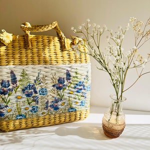 Puede incluir: Un bolso de paja tejido con estampado floral y ribete blanco. El bolso tiene un asa superior y est&aacute; sobre una superficie blanca junto a un jarr&oacute;n de vidrio transparente con flores blancas.