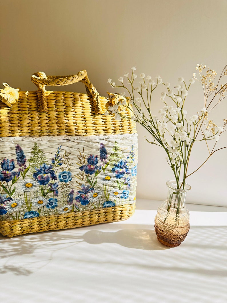 Puede incluir: Un bolso de paja tejido con estampado floral y ribete blanco. El bolso tiene un asa superior y est&aacute; sobre una superficie blanca junto a un jarr&oacute;n de vidrio transparente con flores blancas.