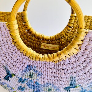May include: A half-moon shaped straw bag with a light purple and yellow woven design. The bag has a wooden handle and a floral print with hummingbirds. The bag has a small tag that reads "Hands of Namaste".