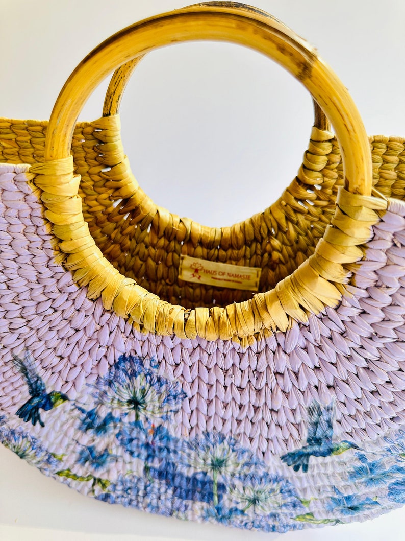 May include: A half-moon shaped straw bag with a light purple and yellow woven design. The bag has a wooden handle and a floral print with hummingbirds. The bag has a small tag that reads "Hands of Namaste".