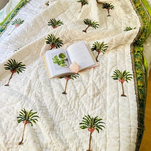 May include: White quilted bedspread with a green and brown border, featuring a repeating pattern of green palm trees with pink accents. An open book and a pink crystal are on the bedspread.
