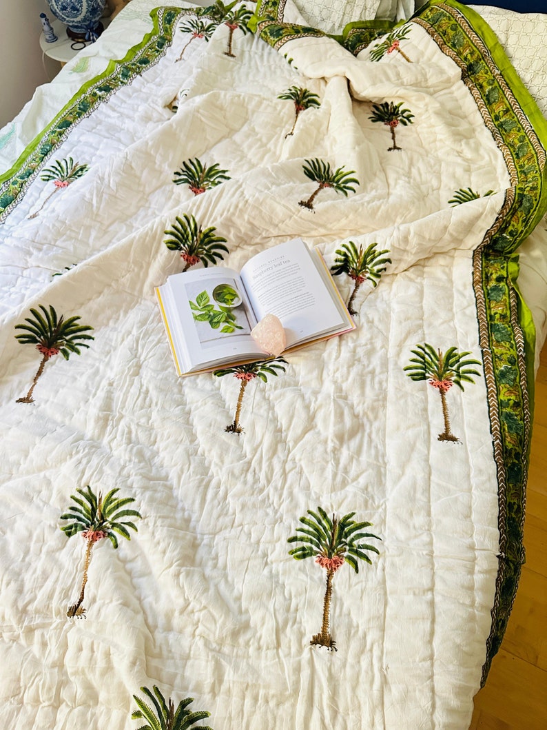 May include: White quilted bedspread with a green and brown border, featuring a repeating pattern of green palm trees with pink accents. An open book and a pink crystal are on the bedspread.