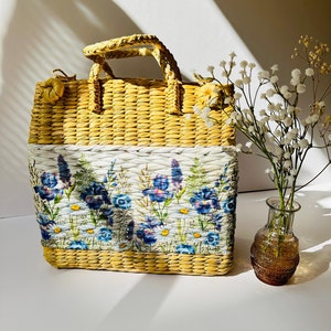 Puede incluir: Un bolso de mano de paja amarilla tejida con un panel de tela blanca con un estampado floral de flores azules y moradas. El bolso tiene un asa tejida y est&aacute; sobre una superficie blanca junto a un jarr&oacute;n de vidrio con flores blancas.