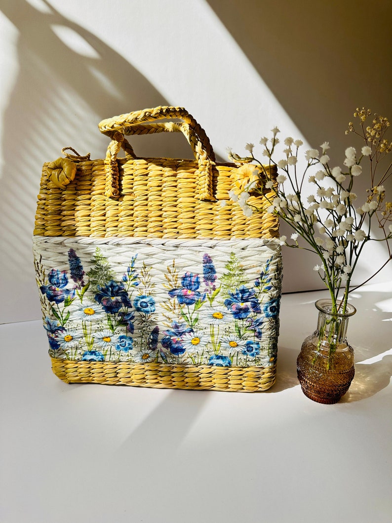 Puede incluir: Un bolso de mano de paja amarilla tejida con un panel de tela blanca con un estampado floral de flores azules y moradas. El bolso tiene un asa tejida y est&aacute; sobre una superficie blanca junto a un jarr&oacute;n de vidrio con flores blancas.