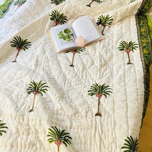 May include: Cream-colored quilt with a repeating pattern of embroidered palm trees in green, brown, and pink. The quilt has a green and gold border. An open book and a pink crystal are on top of the quilt.