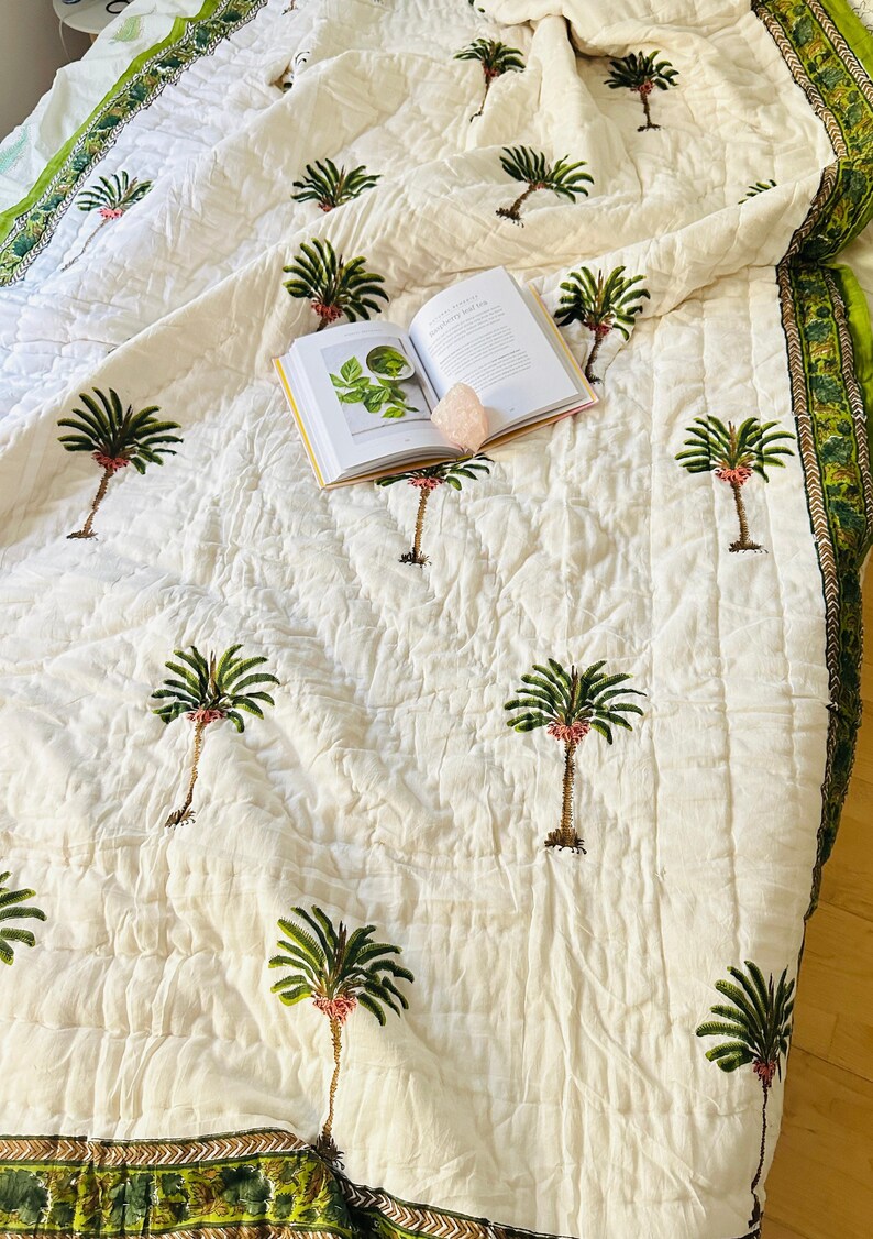 May include: Cream-colored quilt with a repeating pattern of embroidered palm trees in green, brown, and pink. The quilt has a green and gold border. An open book and a pink crystal are on top of the quilt.
