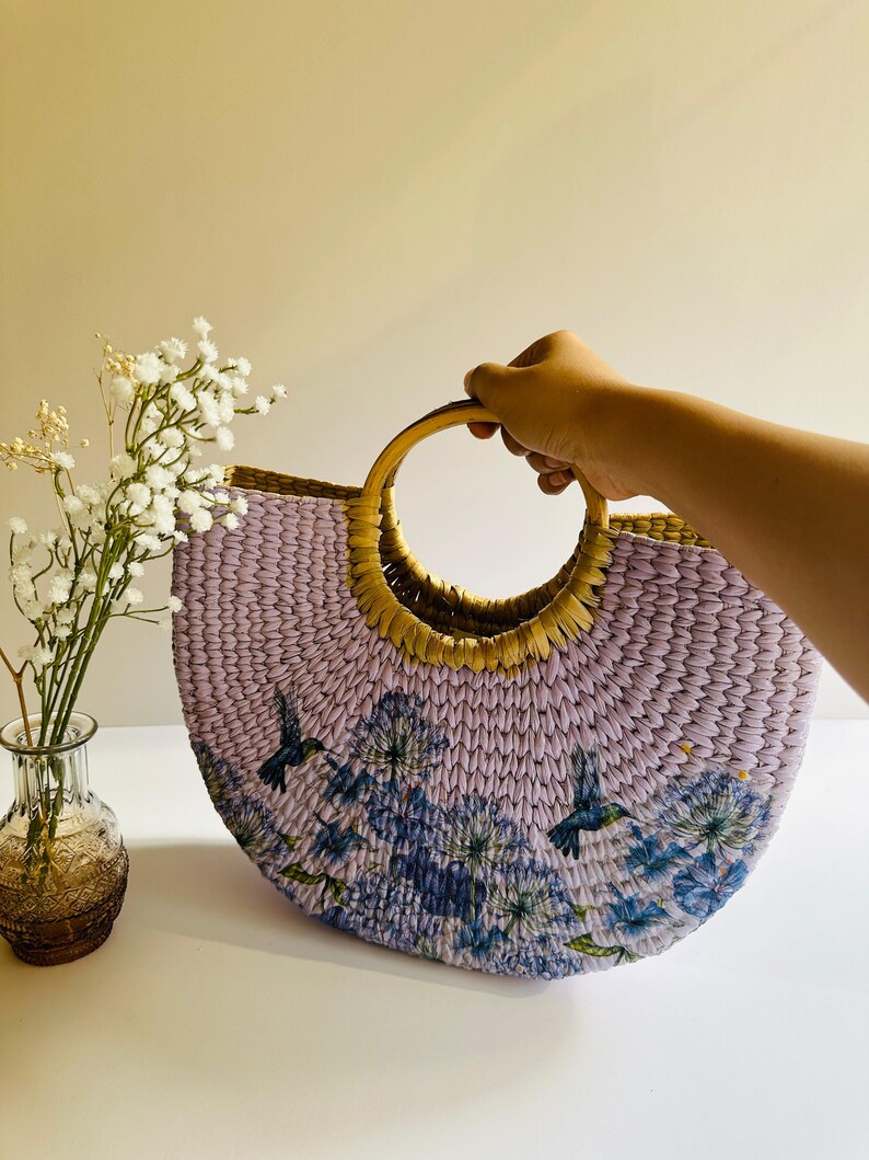 May include: A lavender woven tote bag with a wooden handle. The bag has a floral print with blue hummingbirds.