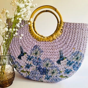 May include: A lavender woven straw tote bag with a double-ringed wooden handle. The bag features a floral print with hummingbirds.
