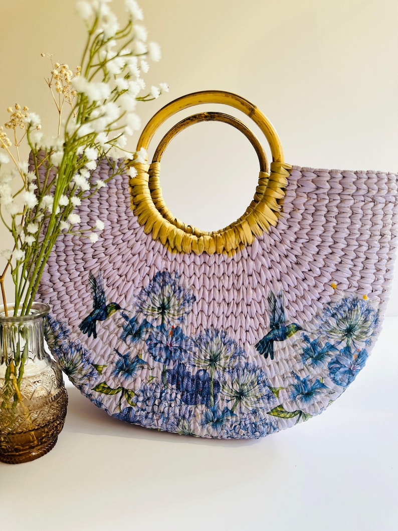 May include: A lavender woven straw tote bag with a double-ringed wooden handle. The bag features a floral print with hummingbirds.