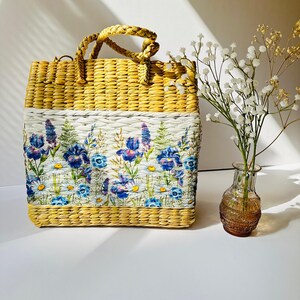 Puede incluir: Un bolso de mano de paja tejido con estampado floral. El bolso tiene un fondo blanco con flores azules y moradas. El bolso tiene un asa tejida marr&oacute;n.