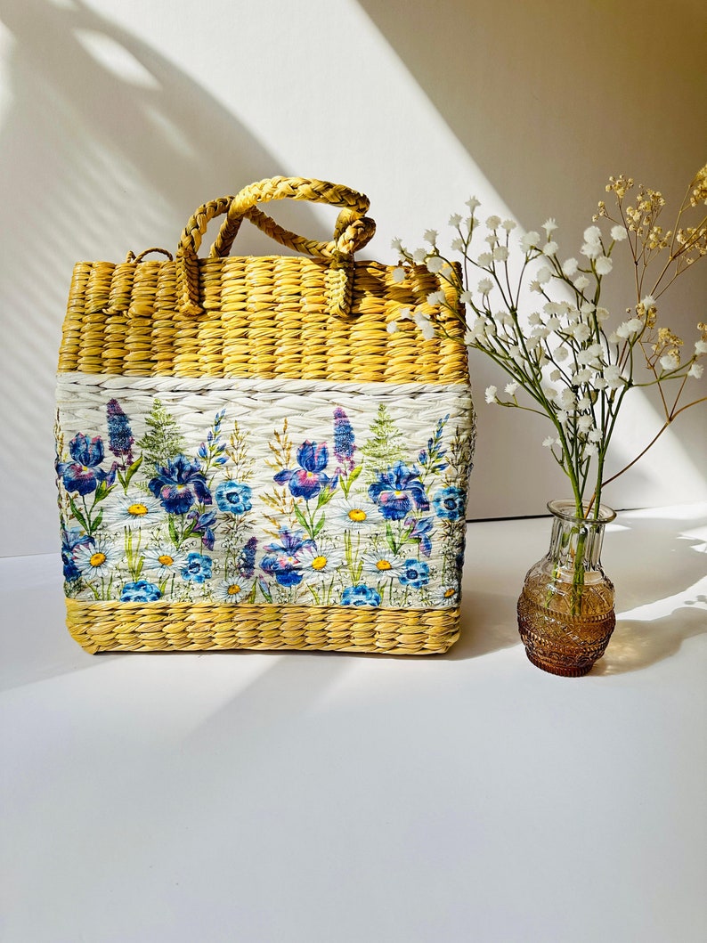Puede incluir: Un bolso de mano de paja tejido con estampado floral. El bolso tiene un fondo blanco con flores azules y moradas. El bolso tiene un asa tejida marr&oacute;n.
