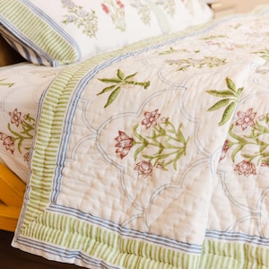 Può includere: Set di trapunte e cuscini floreali bianchi con bordi a righe verdi e blu. La biancheria da letto presenta un motivo ripetuto di fiori e foglie rosse e verdi. La trapunta è su un telaio del letto in legno.