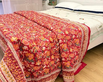 May include: A quilted bedspread featuring a vibrant red base with an intricate floral pattern in shades of orange, yellow, and white. The fabric displays a detailed botanical print with a decorative border, draped over a white bed frame.