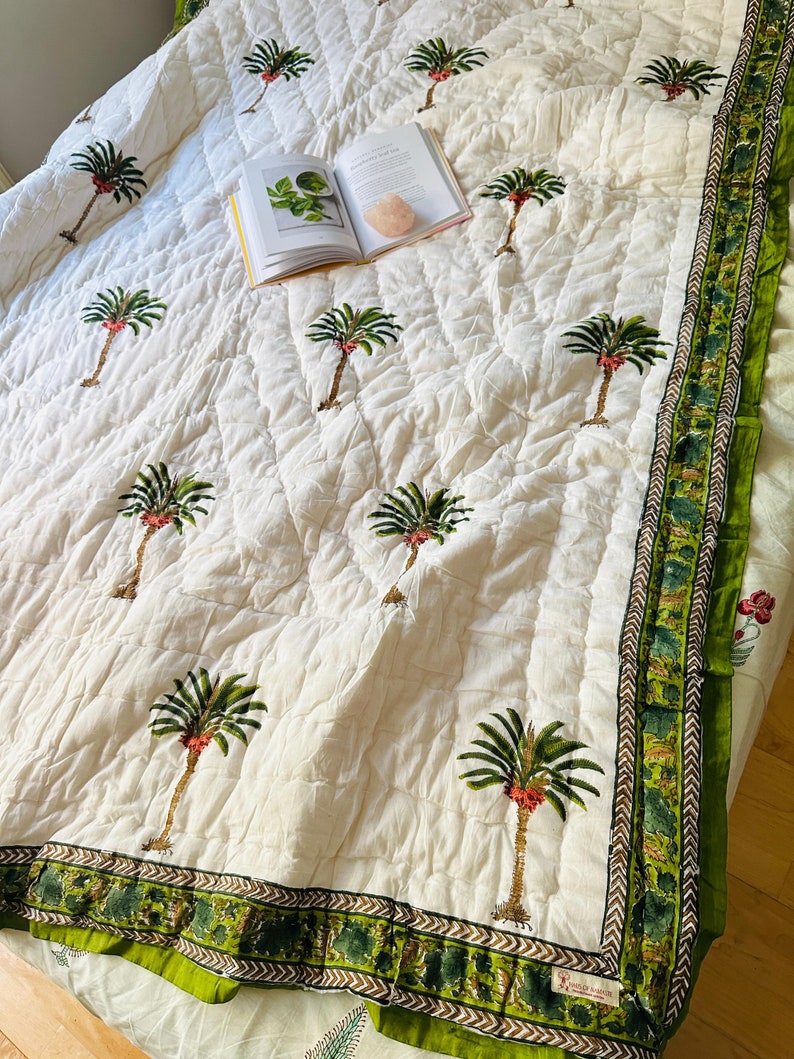 May include: Cream-colored quilted bedspread with a repeating pattern of green palm trees. The border features a green and brown design. An open book and a pink crystal rest on the bedspread.