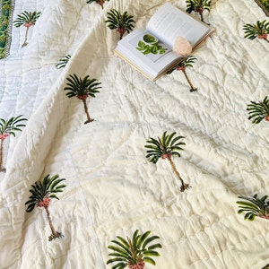 May include: A white quilted bedspread with a repeating pattern of green palm trees and a green and gold border. An open book and a pink crystal are on the bedspread.