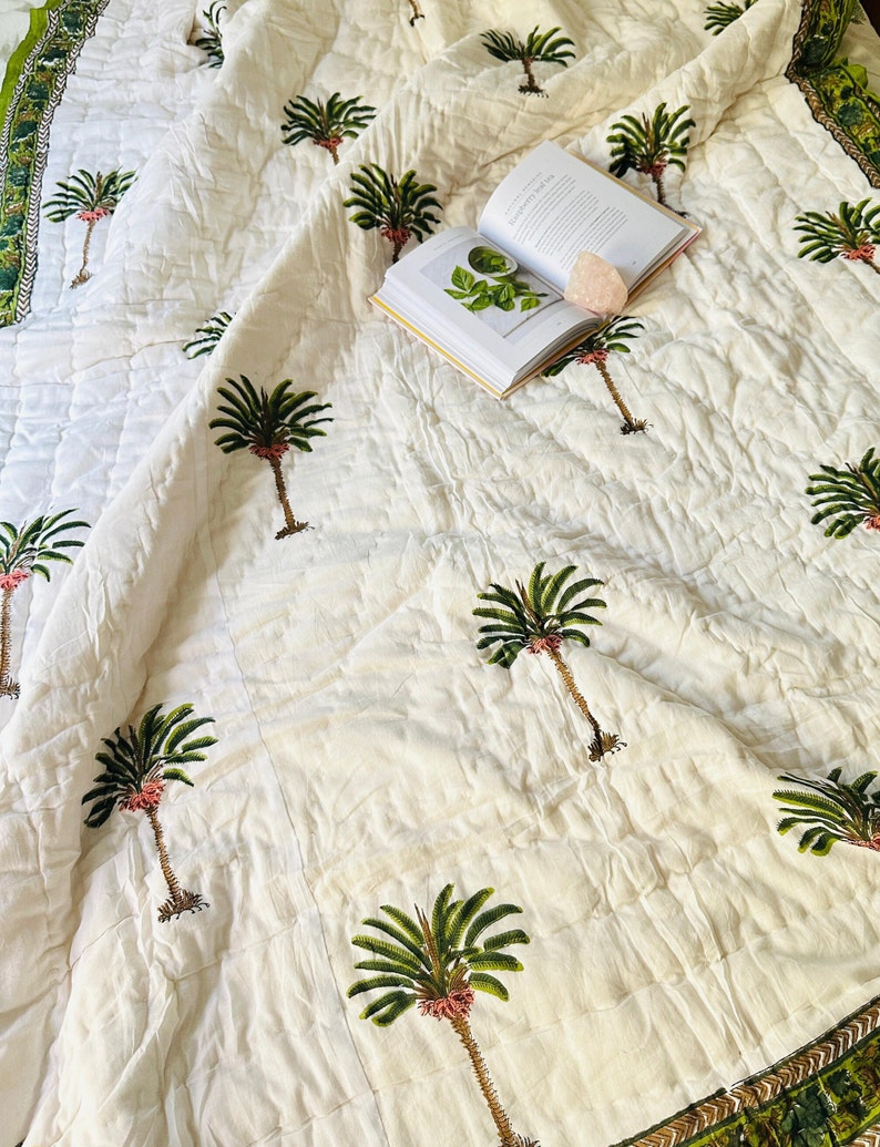 May include: A white quilted bedspread with a repeating pattern of green palm trees and a green and gold border. An open book and a pink crystal are on the bedspread.