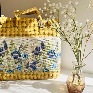 Puede incluir: Un bolso de paja tejido amarillo con un forro de estampado floral. El bolso tiene un asa superior y est&aacute; sentado sobre una superficie blanca junto a un jarr&oacute;n de vidrio transparente con flores blancas.