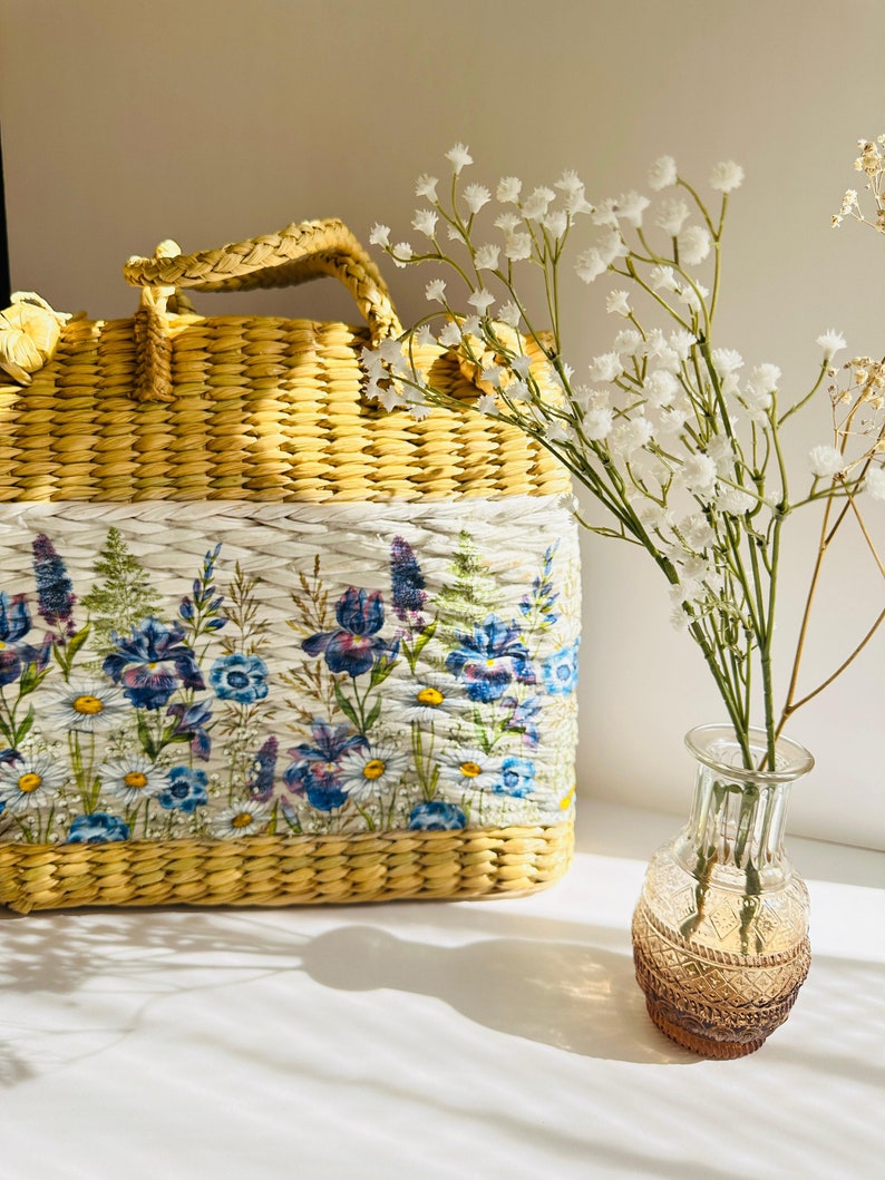 Puede incluir: Un bolso de paja tejido amarillo con un forro de estampado floral. El bolso tiene un asa superior y est&aacute; sentado sobre una superficie blanca junto a un jarr&oacute;n de vidrio transparente con flores blancas.
