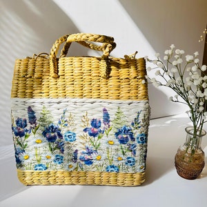 Puede incluir: Un bolso de mano de paja tejido con estampado floral. El estampado presenta iris azules y morados, margaritas blancas y hojas verdes sobre un fondo blanco.