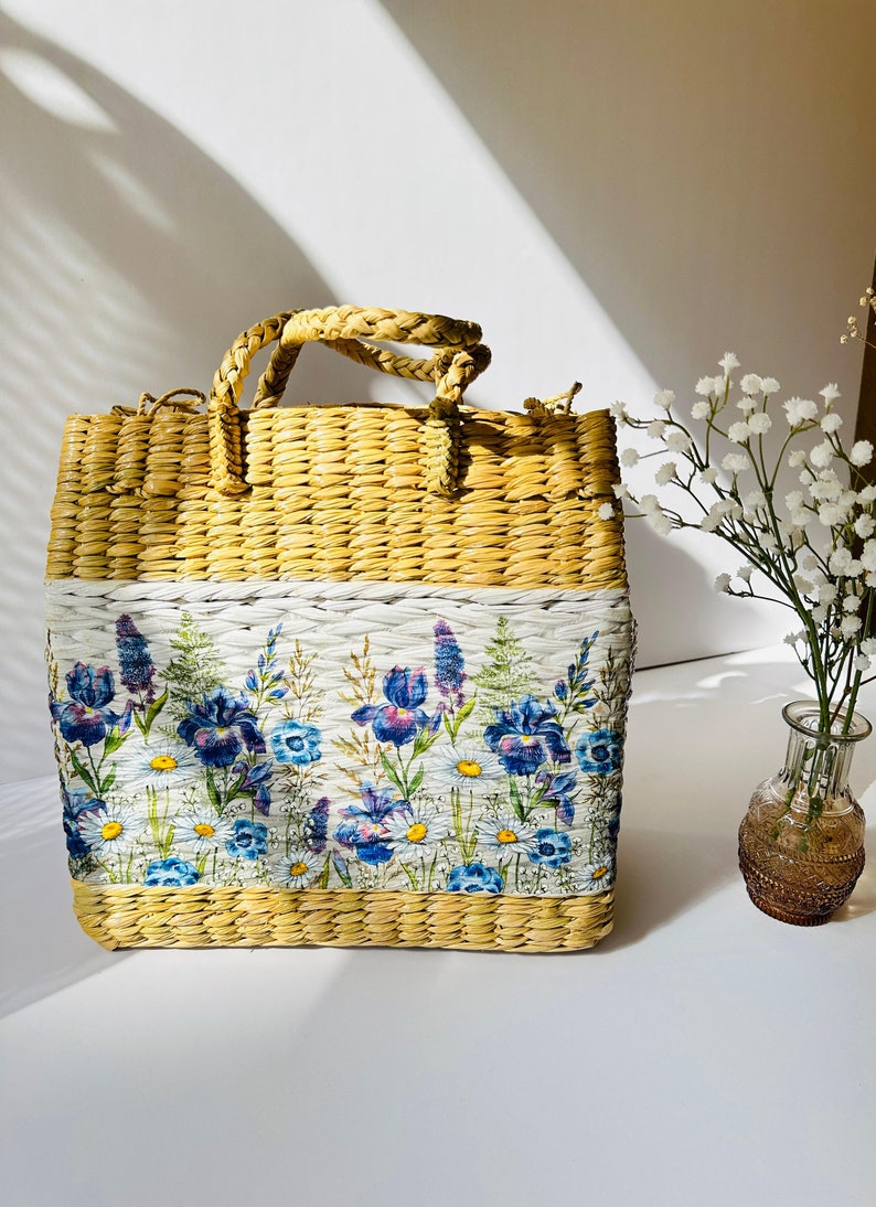 Puede incluir: Un bolso de mano de paja tejido con estampado floral. El estampado presenta iris azules y morados, margaritas blancas y hojas verdes sobre un fondo blanco.