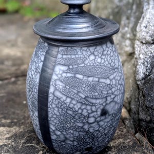 May include: A ceramic jar with a lid featuring a black and white crackle glaze finish. The vessel has a rounded body with a smooth, solid black vertical stripe running down the side, contrasting with the textured, web-like pattern across the surface.