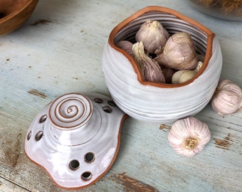 May include: White ceramic garlic keeper with a lid. The keeper has a textured, wavy design and is filled with garlic cloves. The lid has a spiral design and holes for ventilation.
