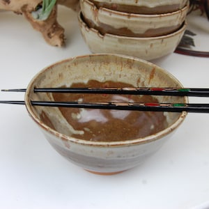 May include: A brown and white ceramic bowl with a pair of black chopsticks resting on the rim. The bowl has a rustic, handmade look.
